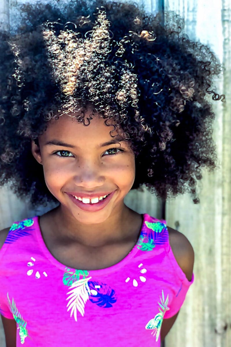 A joyful young girl showcases her beautiful curly Afro hair and bright smile in an outdoor portrait against a wooden backdrop.